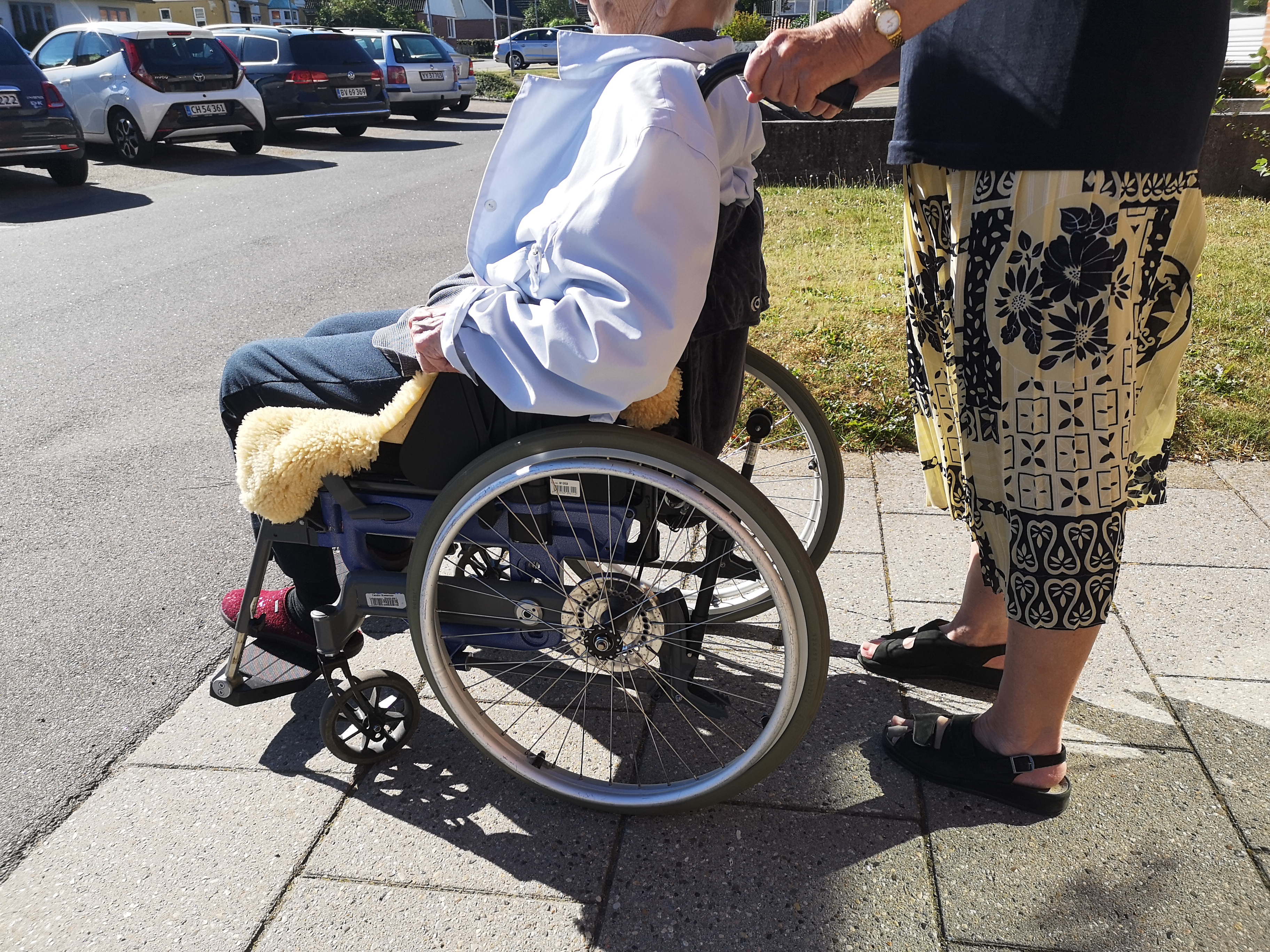 Nærbillede af to personer, ud mod en vej, hvoraf den ene sidder i kørestol.