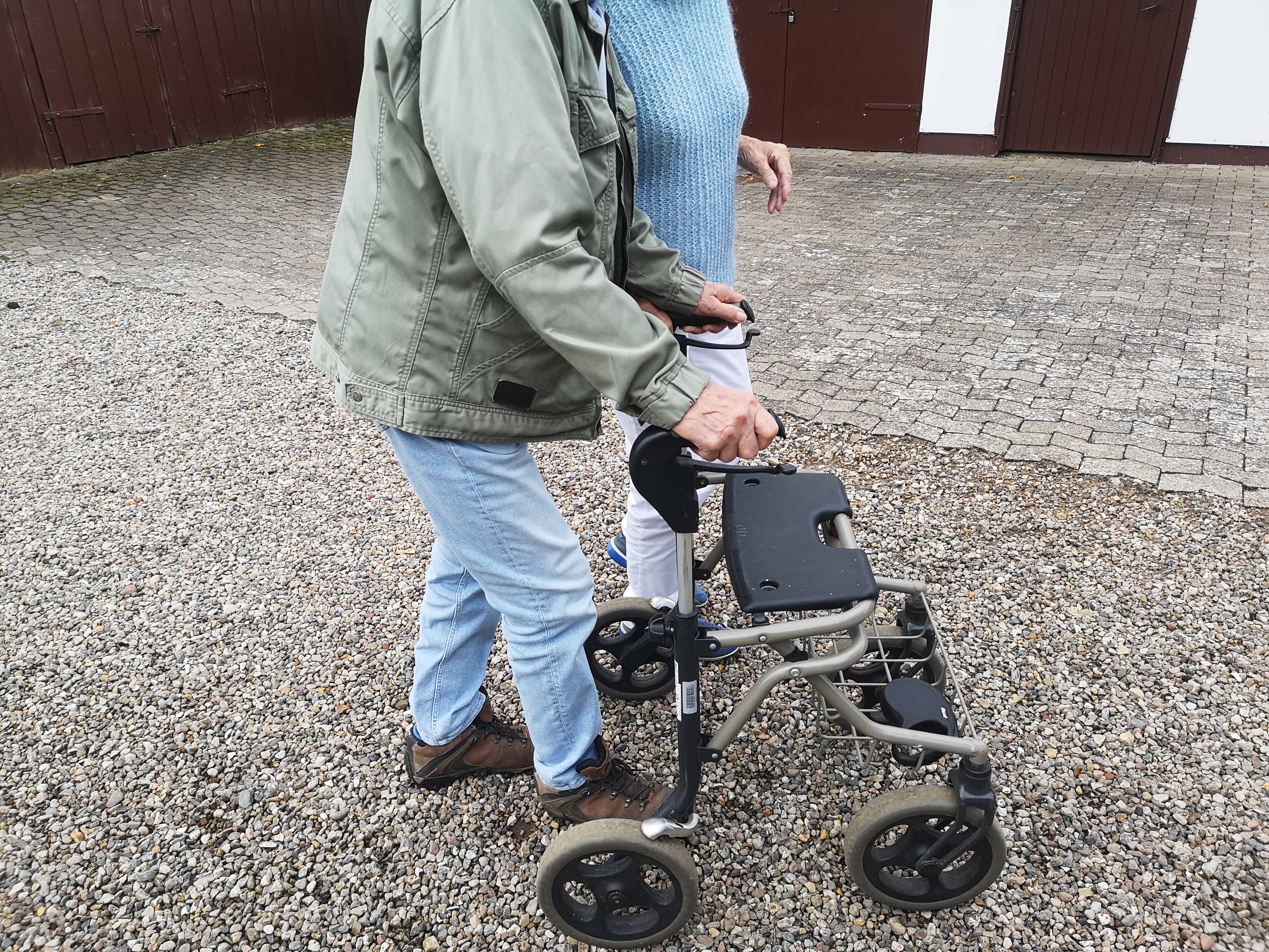 Et nærbillede af to personer, der går på en gårdsplads, hvoraf den ene går med rollator.