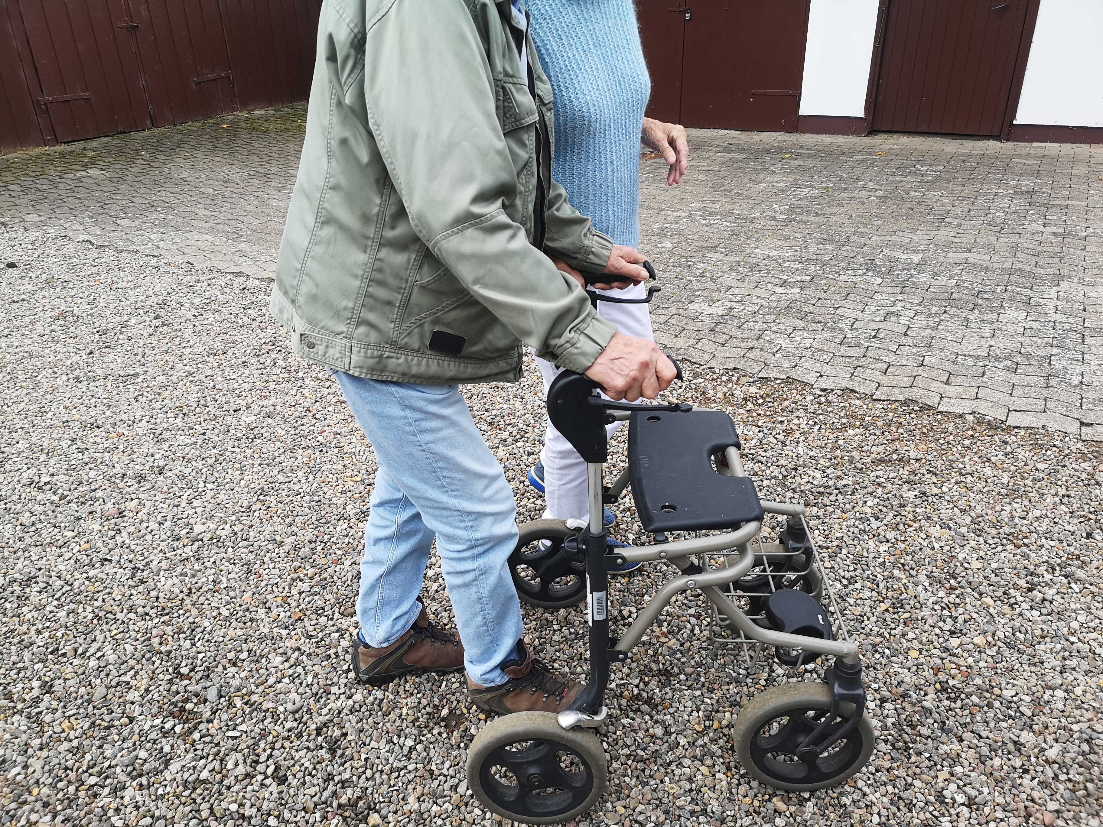 Et nærbillede af to personer, der går på en gårdsplads, hvoraf den ene går med rollator.